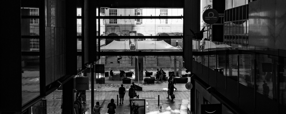 A grey scale image of an office space, looking out the window at workers walking past.