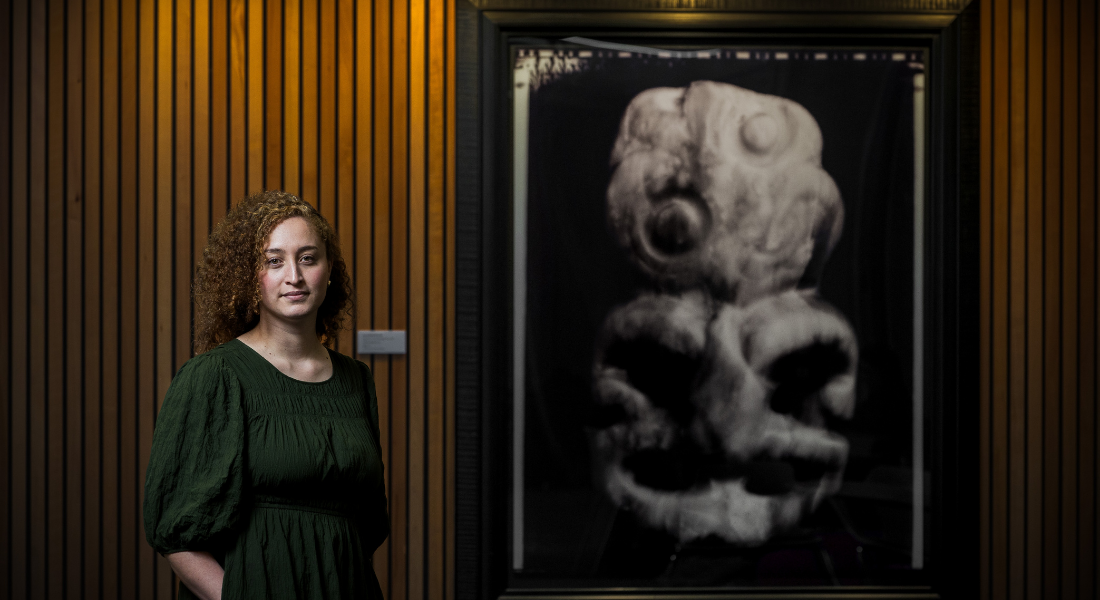 Maōri wahine, long curly dark hair, green dress, standing in front of wooden wall beside large photograph of a tiki.