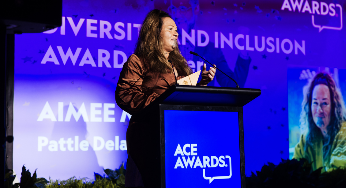 Māori wāhine, receiving award on stage, wearing brown silk dress, long hair.