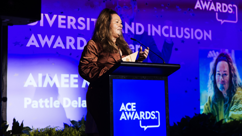 Maōri wāhine, awards evening, on stage, wearing brown silk dress, long hair, moko, holding award.
