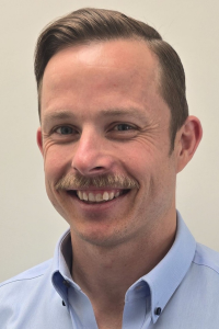 White male, dark brown hair, moustache, smiling, blue shirt, headshot.