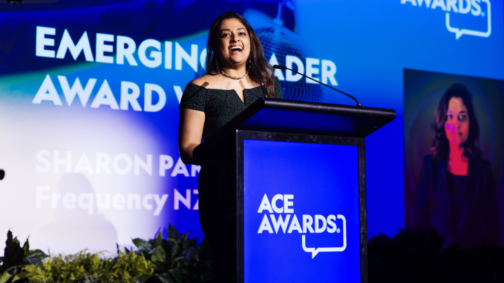 Dark skinned woman, long hair, smiling, evening dress, awards ceremony, standing at podium receiving award.