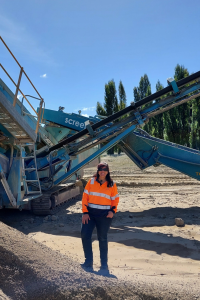 White female, smiling, high vis, on site, machinery in background, sunny day.