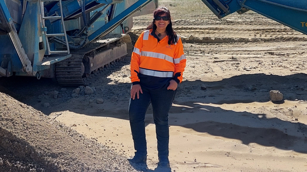 Female, long dark hair, sunglasses, smiling, wearing high vis, standing at construction site.